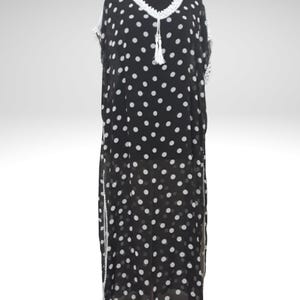 May include: Black and white polka dot dress with white crochet trim and a tassel. The dress is displayed on a mannequin. The dress has a V-neckline and a loose, flowing silhouette. The dress is floor-length.