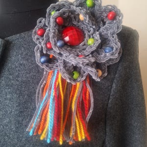 May include: A gray crocheted flower brooch with a red jewel in the center. The flower is decorated with colorful wooden beads and has a long fringe of colorful yarn.