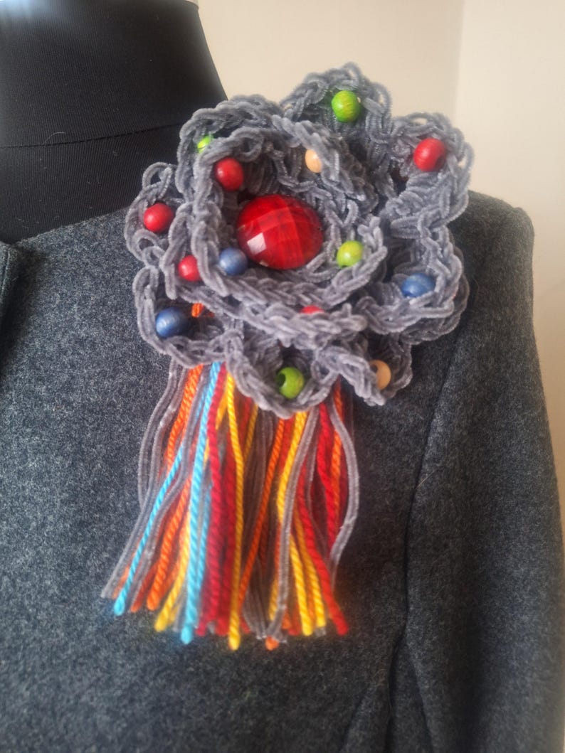 May include: A gray crocheted flower brooch with a red jewel in the center. The flower is decorated with colorful wooden beads and has a long fringe of colorful yarn.