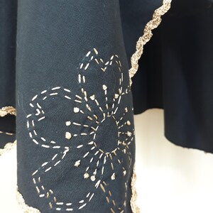 A close-up of the bottom edge of a black linen garment, showing gold trim and a gold embroidered flower design.
