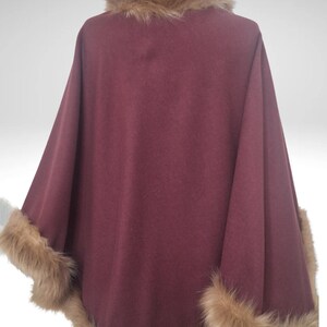 May include: A burgundy cape with faux fur trim on the edges and collar. The cape is displayed on a mannequin, highlighting its flowing design and luxurious texture. The fur trim is a light brown colour.
