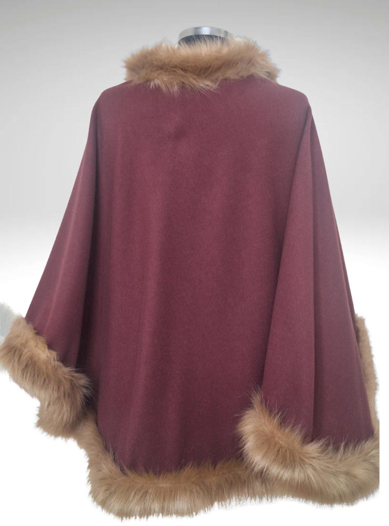 May include: A burgundy cape with faux fur trim on the edges and collar. The cape is displayed on a mannequin, highlighting its flowing design and luxurious texture. The fur trim is a light brown colour.