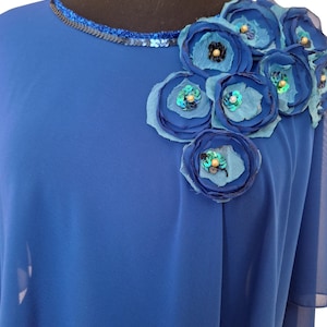 May include: Royal blue top with a round neckline trimmed with sequins. The shoulder is adorned with fabric flowers in shades of blue, embellished with sequins and beads. The top has a flowing, draped design.