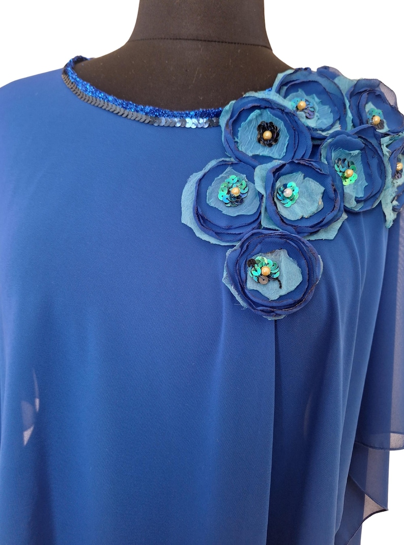 May include: Royal blue top with a round neckline trimmed with sequins. The shoulder is adorned with fabric flowers in shades of blue, embellished with sequins and beads. The top has a flowing, draped design.
