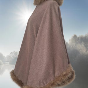 the reversible poncho with a faux fur trim around the neckline and hem, made of a soft, cashmere fabric and has a relaxed fit.  It is shown on a mannequin against a blurry background of a lake and trees. The fur is a light brown color, complementing the overall muted tone of the garment.