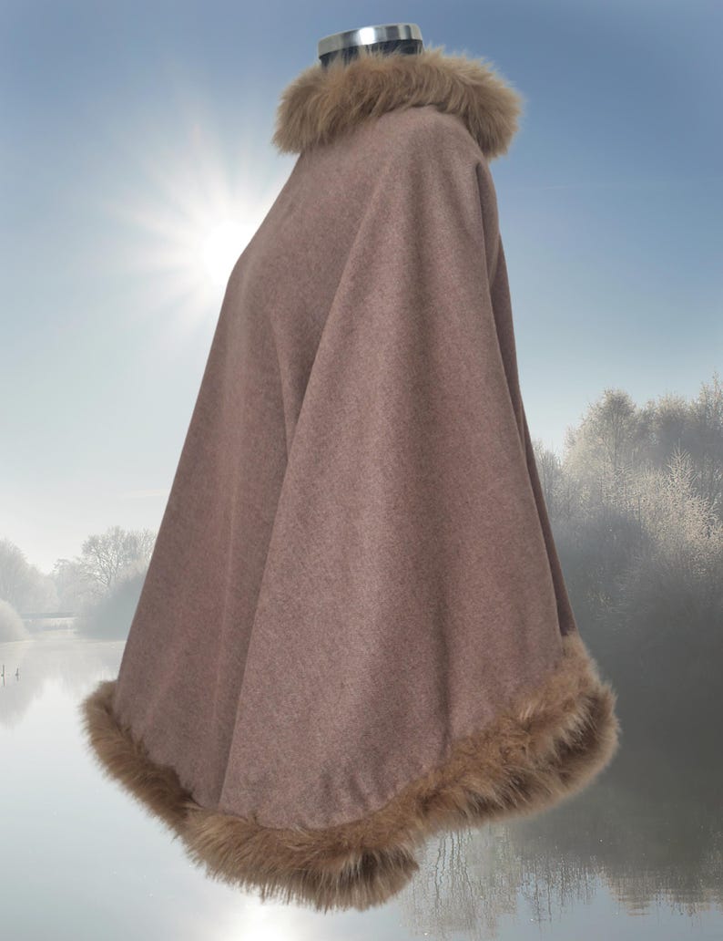 the reversible poncho with a faux fur trim around the neckline and hem, made of a soft, cashmere fabric and has a relaxed fit.  It is shown on a mannequin against a blurry background of a lake and trees. The fur is a light brown color, complementing the overall muted tone of the garment.