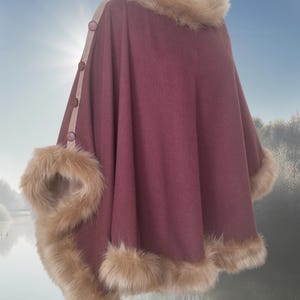 A maroon poncho with beige faux fur trim around the neckline and hem.  The poncho features a beige accent panel along the sleeves with brown buttons.  The garment is shown on a mannequin against a blurry background of a lake and trees.