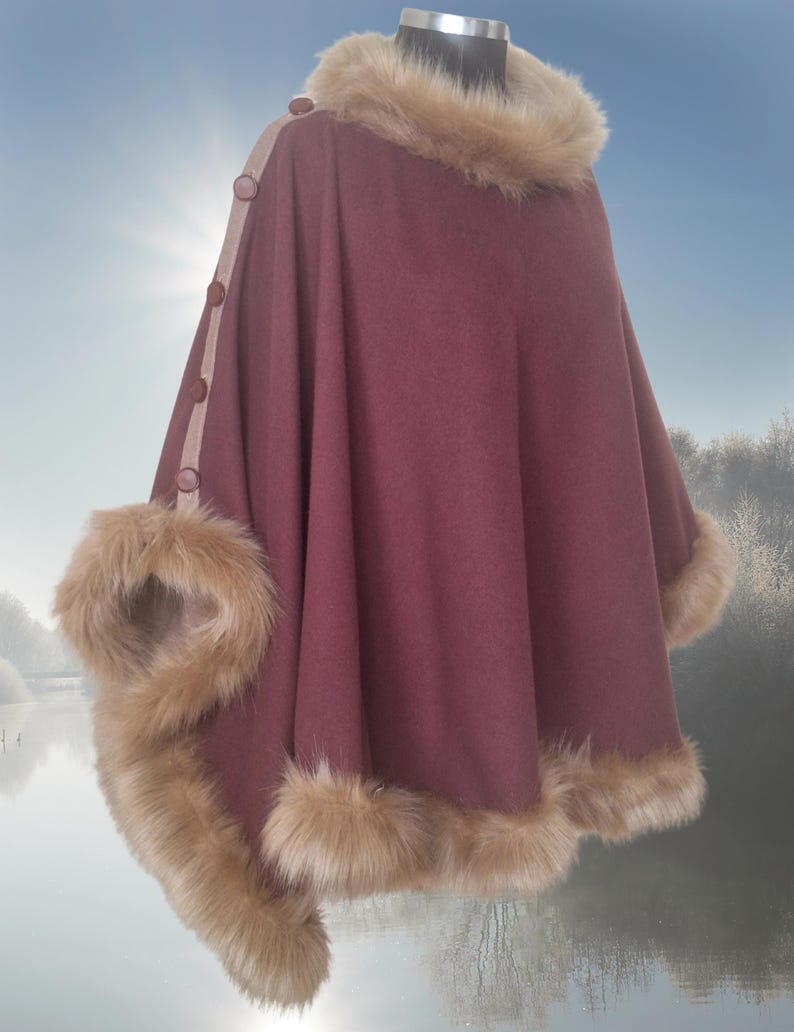 A maroon poncho with beige faux fur trim around the neckline and hem.  The poncho features a beige accent panel along the sleeves with brown buttons.  The garment is shown on a mannequin against a blurry background of a lake and trees.