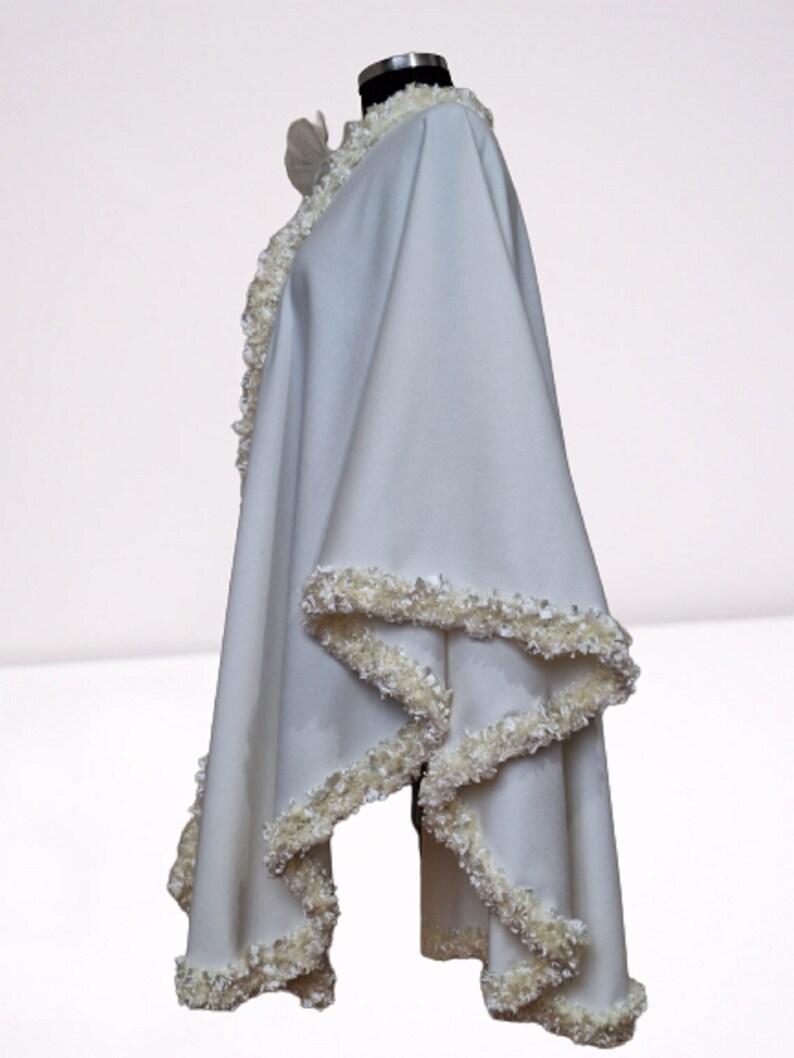 Side view of an off-white cashmere wedding cape showcasing the draped flow of the fabric and the textured handmade trim along the edges. Elegant, vintage-inspired bridal shawl displayed on a mannequin.