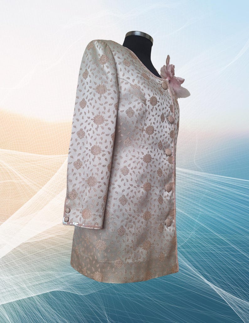 May include: A long-sleeved, cream-colored jacket with a floral pattern. The jacket has a button closure and a pink flower embellishment on the collar.