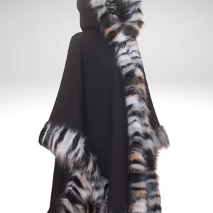 May include: Black hooded cape with faux fur trim. The cape features a hood and is trimmed with a striped pattern of black, white, and tan faux fur along the hood, sleeves, and hemline. The cape is made of a soft, dark fabric.