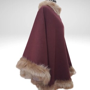 May include: A burgundy cape with a faux fur collar and trim. The cape is displayed on a mannequin. The fur trim is a light brown colour, adding a touch of luxury to the garment. The cape's design is flowing.