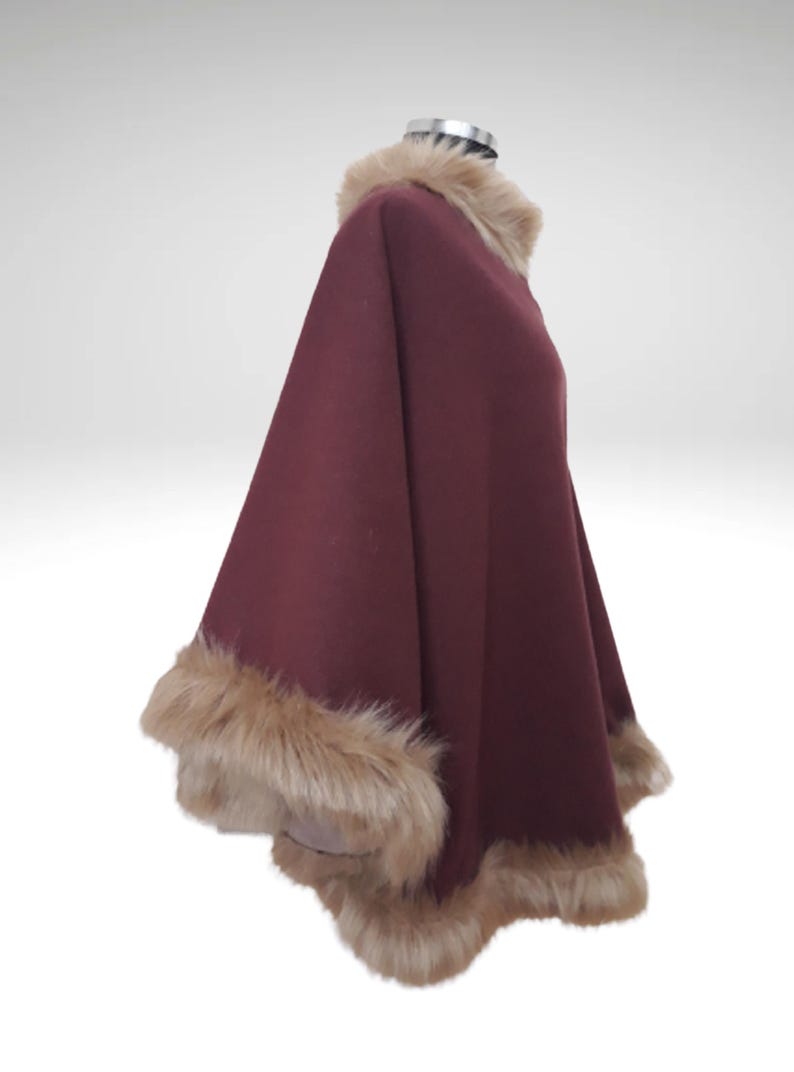 May include: A burgundy cape with a faux fur collar and trim. The cape is displayed on a mannequin. The fur trim is a light brown colour, adding a touch of luxury to the garment. The cape's design is flowing.