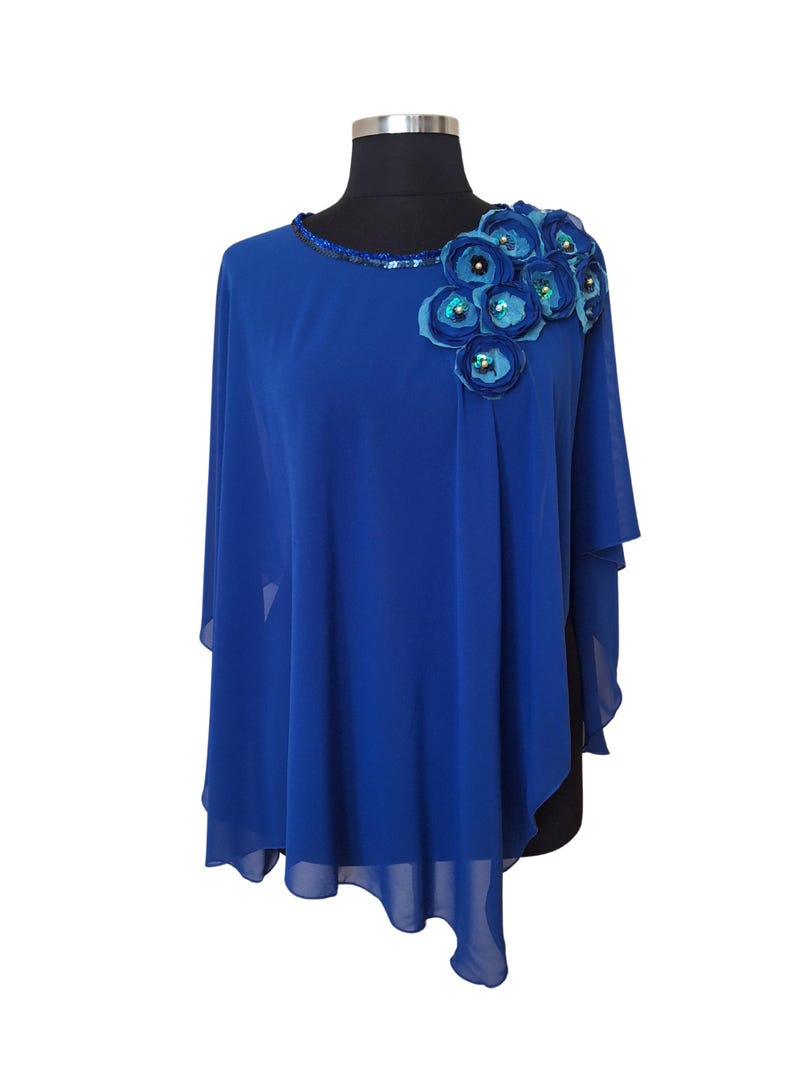 May include: A royal blue cape-style top with a round neckline and a cluster of blue fabric flowers with sequin centres on the shoulder. The neckline is embellished with sequins. The top is displayed on a mannequin.