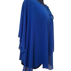 May include: A royal blue cape-style top with a decorative floral embellishment on the shoulder. The top is made of a flowing, sheer fabric and features a round neckline with beaded trim. The garment is displayed on a black mannequin.