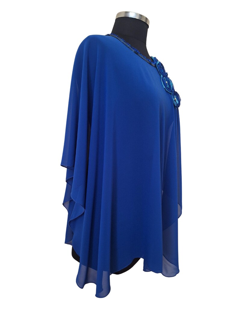 May include: A royal blue cape-style top with a decorative floral embellishment on the shoulder. The top is made of a flowing, sheer fabric and features a round neckline with beaded trim. The garment is displayed on a black mannequin.