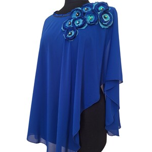May include: A royal blue cape-style top with a floral embellishment on the shoulder. The flowers are in shades of blue and adorned with sequins. The neckline is embellished with small beads.