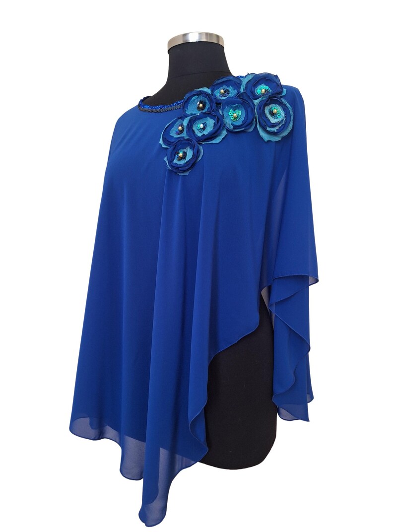 May include: A royal blue cape-style top with a floral embellishment on the shoulder. The flowers are in shades of blue and adorned with sequins. The neckline is embellished with small beads.