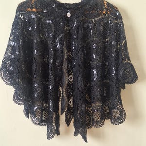 May include: Black lace capelet with sequin accents, hanging on a wooden hanger. The capelet features a round neckline, short sleeves, and a button closure. The intricate lace design is visible throughout the garment.