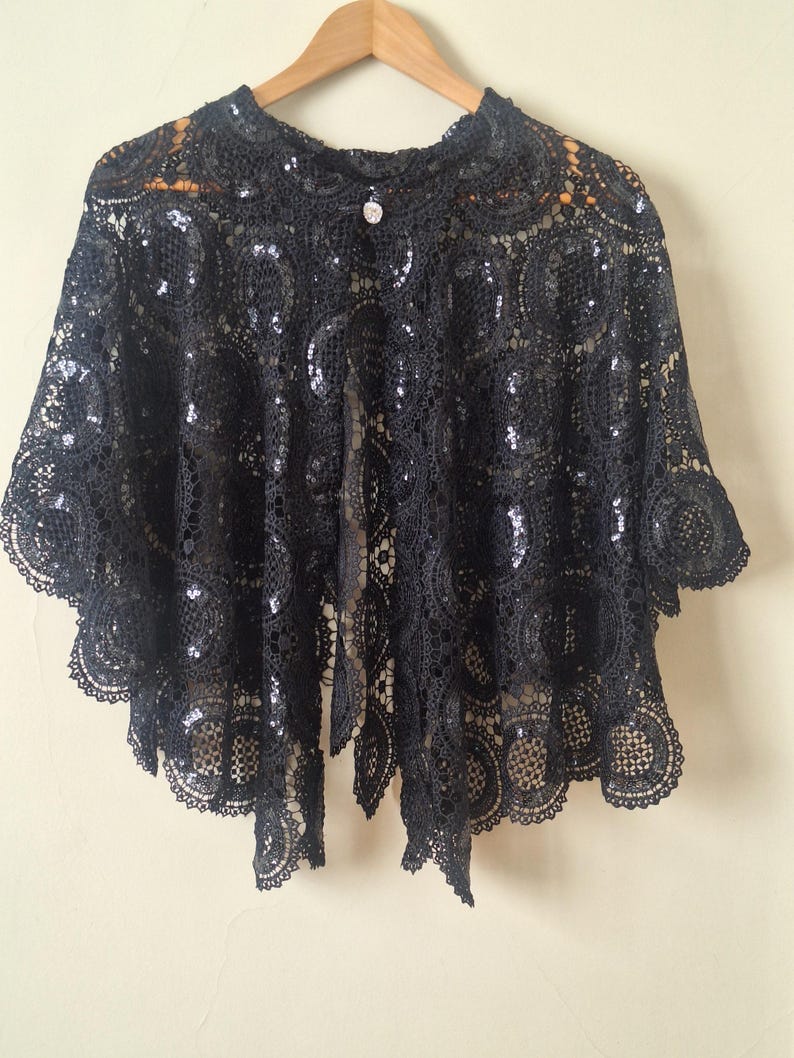 May include: Black lace capelet with sequin accents, hanging on a wooden hanger. The capelet features a round neckline, short sleeves, and a button closure. The intricate lace design is visible throughout the garment.