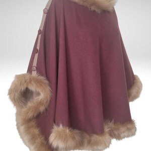May include: A burgundy cape with faux fur trim and collar. The cape has a button closure and a tan trim along the front. The faux fur is light brown. This outerwear garment is designed for warmth and style.