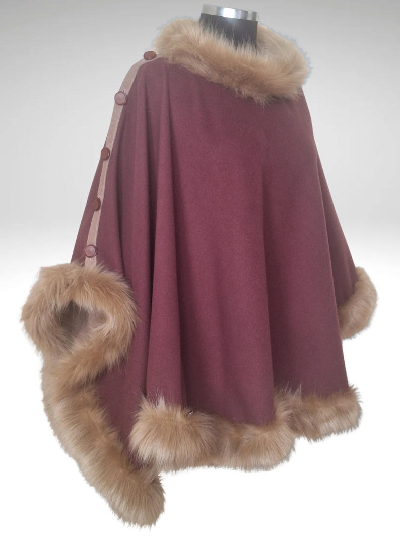 May include: A burgundy cape with faux fur trim and collar. The cape has a button closure and a tan trim along the front. The faux fur is light brown. This outerwear garment is designed for warmth and style.