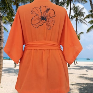 Puede incluir: Batista naranja con un dise&ntilde;o de flor de hibisco negro en la espalda. La bata tiene un cintur&oacute;n de atar y est&aacute; hecha de una tela ligera.