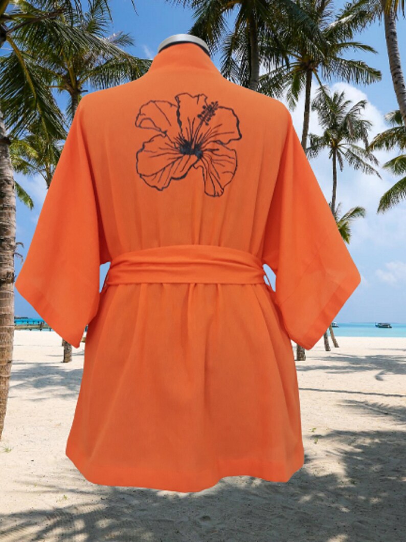 Puede incluir: Batista naranja con un dise&ntilde;o de flor de hibisco negro en la espalda. La bata tiene un cintur&oacute;n de atar y est&aacute; hecha de una tela ligera.