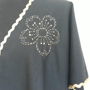 A black linen garment with gold trim and a large, gold embroidered flower design on the shoulder, displayed on a black mannequin.