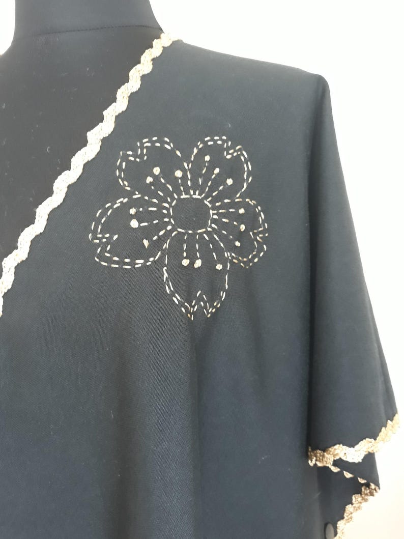 A black linen garment with gold trim and a large, gold embroidered flower design on the shoulder, displayed on a black mannequin.