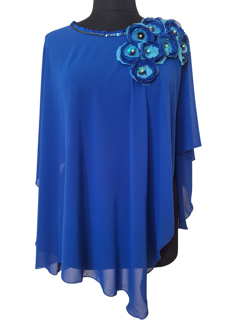 May include: A royal blue cape-style top with a round neckline embellished with sequins. The shoulder is adorned with fabric flowers in shades of blue and teal, accented with beads. The cape has a flowing, asymmetrical hemline.