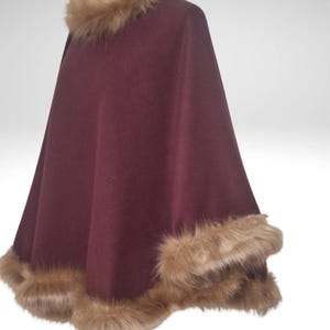 May include: A claret-coloured cape with a faux fur collar and trim. The cape is made of a soft material and has a flowing design. The faux fur is a light brown colour, adding a touch of luxury to the garment.