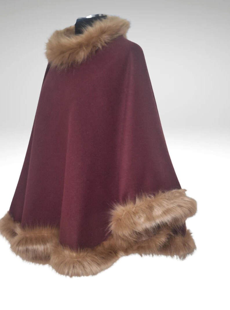 May include: A claret-coloured cape with a faux fur collar and trim. The cape is made of a soft material and has a flowing design. The faux fur is a light brown colour, adding a touch of luxury to the garment.