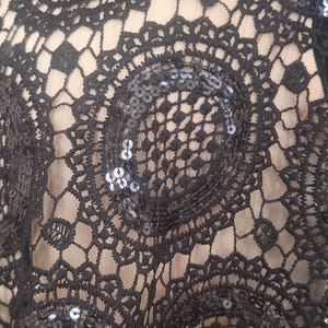May include: Close-up of black lace fabric with a circular, openwork design. The lace is embellished with small, shiny sequins, adding a touch of sparkle. The background is a light beige color, providing contrast to the dark lace.
