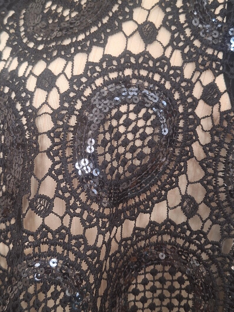 May include: Close-up of black lace fabric with a circular, openwork design. The lace is embellished with small, shiny sequins, adding a touch of sparkle. The background is a light beige color, providing contrast to the dark lace.