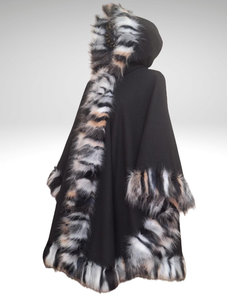 May include: A black hooded cape with faux fur trim. The cape features a hood and is trimmed with a mix of black, white, and tan faux fur along the edges and hood. The cape is designed to be worn over the shoulders.