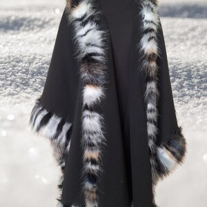 May include: A black cape with a faux fur trim. The trim features a mix of white, black, and brown colors. The cape is displayed on a mannequin against a snowy background.