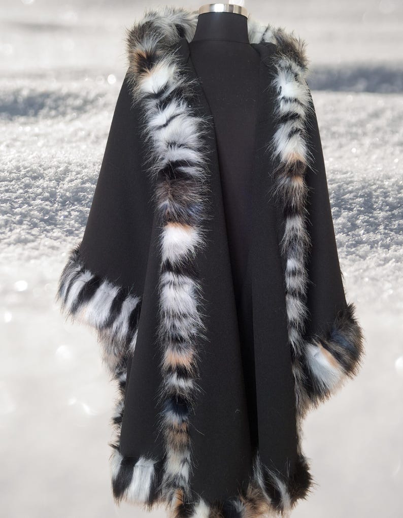 May include: A black cape with a faux fur trim. The trim features a mix of white, black, and brown colors. The cape is displayed on a mannequin against a snowy background.