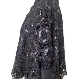 May include: Black lace cape with circular patterns and sequin accents. The cape is displayed on a black mannequin. The intricate design and embellishments create a vintage or formal aesthetic. The garment is likely for special occasions.