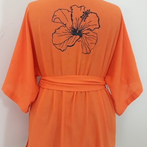 Puede incluir: Bata de color naranja con una flor de hibisco negra bordada en la espalda. La bata tiene un cintur&oacute;n de atar.