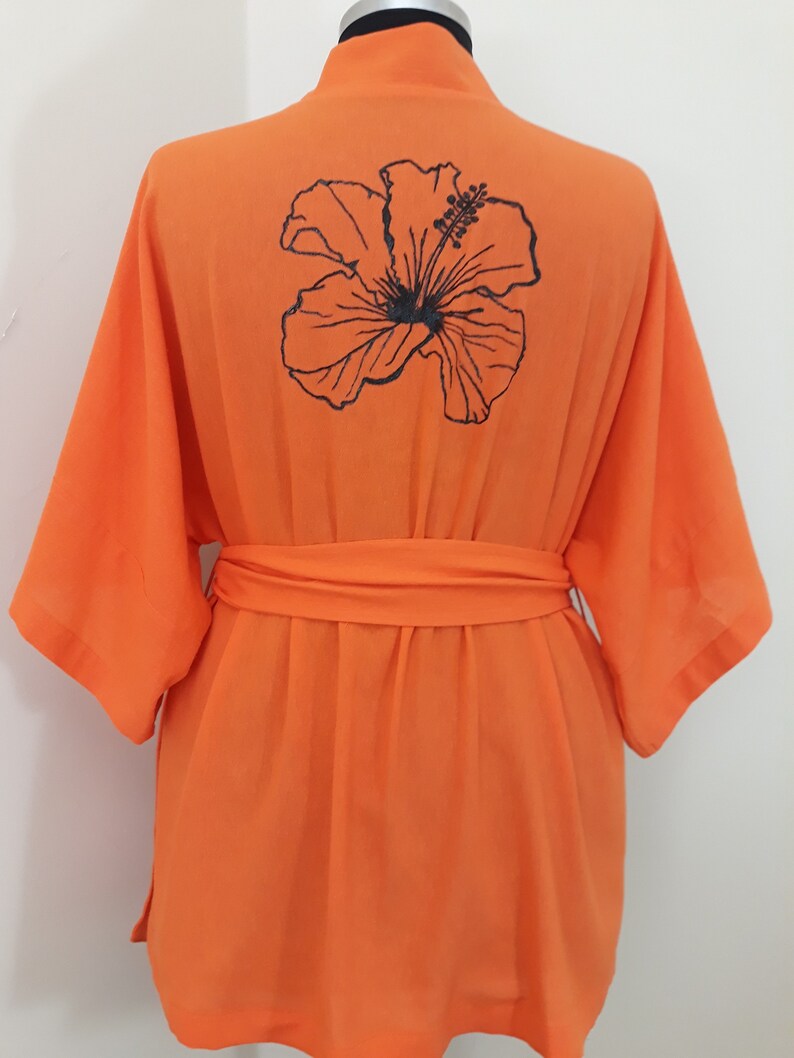 Puede incluir: Bata de color naranja con una flor de hibisco negra bordada en la espalda. La bata tiene un cintur&oacute;n de atar.