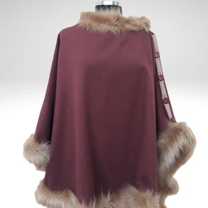 May include: A burgundy cape with faux fur trim on the collar and cuffs. The cape has a button closure on the right shoulder and a decorative trim. The faux fur is light brown, and the cape is displayed on a mannequin.