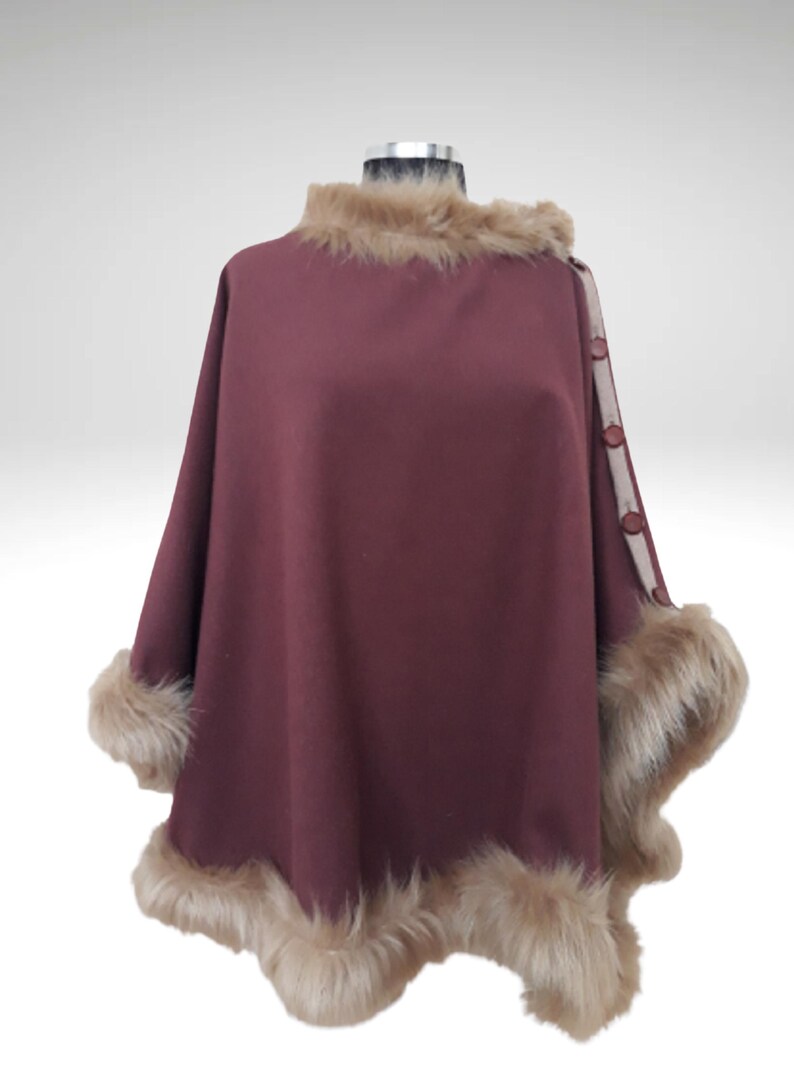 May include: A burgundy cape with faux fur trim on the collar and cuffs. The cape has a button closure on the right shoulder and a decorative trim. The faux fur is light brown, and the cape is displayed on a mannequin.
