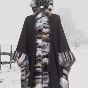 Front side view of black hooded cape featuring dense, long-pile  fur trim in black, white, and caramel hues — luxury handmade winter fashion