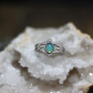 May include: A silver ring with an oval, iridescent, blue-green stone. The ring has a detailed, openwork design with small embellishments. The ring is displayed on a white, textured surface.