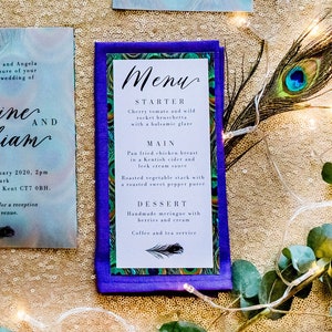 Könnte beinhalten: Eine Hochzeitseinladungssuite mit einem Pfauenfeder-Motiv. Die Einladung ist auf einem Aquarellhintergrund mit den Namen Catherine und William gedruckt. Die RSVP-Karte ist weiß mit einem violetten Rand. Die Speisekarte ist violett mit einem Pfauenfeder-Design und dem Text "Menu" in Weiß. Die Karten sind von Pfauenfedern, Blumen und Lichterketten umgeben.