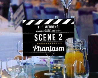 May include: A wedding table setting with a black and white clapperboard table number that reads "The Wedding Starring Kimberly Taylor & Jon Cross Scene 2 Phantasam". The table is set with white linen, silver cutlery, and clear glasses. A jug of orange juice and a bouquet of white and purple flowers are on the table.