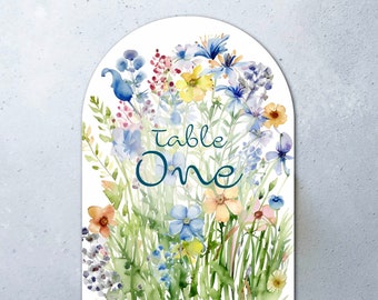 May include: A white card with a watercolor illustration of blue, yellow, and pink flowers. The text "Table One" is written in blue cursive.