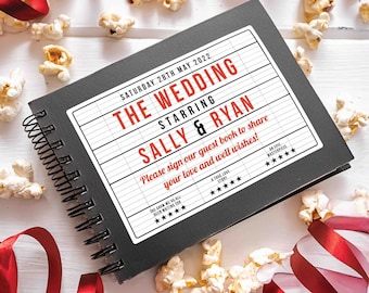 May include: A black guest book with a white label that says "The Wedding" with a movie ticket design. The label also says "Starring Sally & Ryan" and "Please sign our guest book to share your love and well wishes!" The book is surrounded by popcorn and red ribbon.