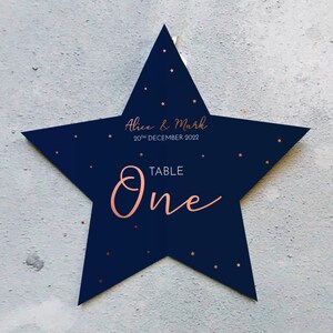 May include: A navy blue star-shaped table sign with copper-colored text and star accents. The sign reads "Alice & Mark 20th December 2022" and "TABLE One". The sign is hanging against a light gray textured wall.